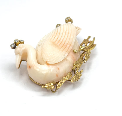 A high three-quarter view from the front, looking down on the carved coral swan's arched neck and feathered back, with the diamond cluster above the head and textured gold reeds fanning out at the side.