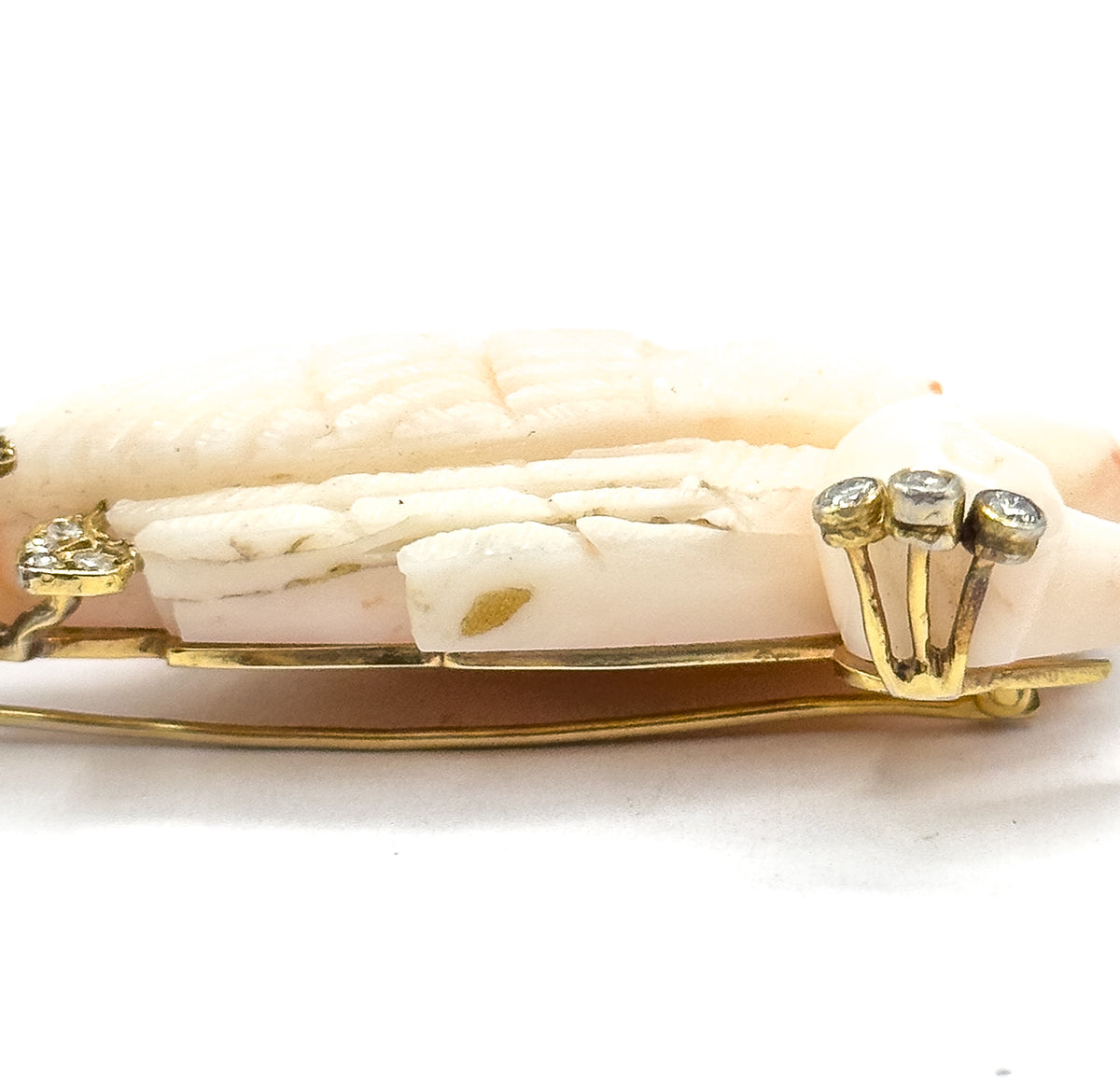 A tight edge-on close-up along the coral's carved feather grooves, with a three-stone diamond cluster raised on prongs at the right and the slim gold pin stem running the full length beneath.