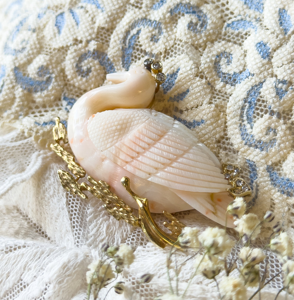 A carved coral swan brooch with diamond accents at the head and back, shown resting on cream lace and blue-patterned fabric, its textured gold reeds curving below the bird's body.