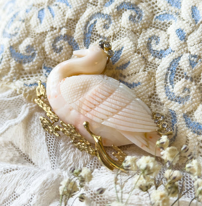 A carved coral swan brooch with diamond accents at the head and back, shown resting on cream lace and blue-patterned fabric, its textured gold reeds curving below the bird's body.