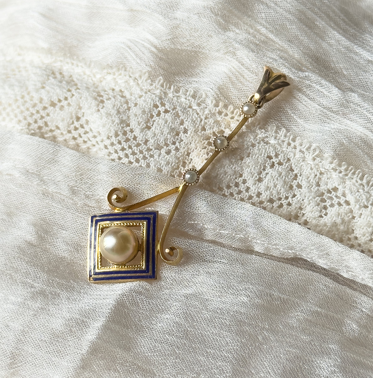 Closer diagonal view showing the three seed pearls spaced along the slender bar and the scrolled gold volutes flanking the cobalt blue enamel square, with the creamy pearl in its milgrain setting.