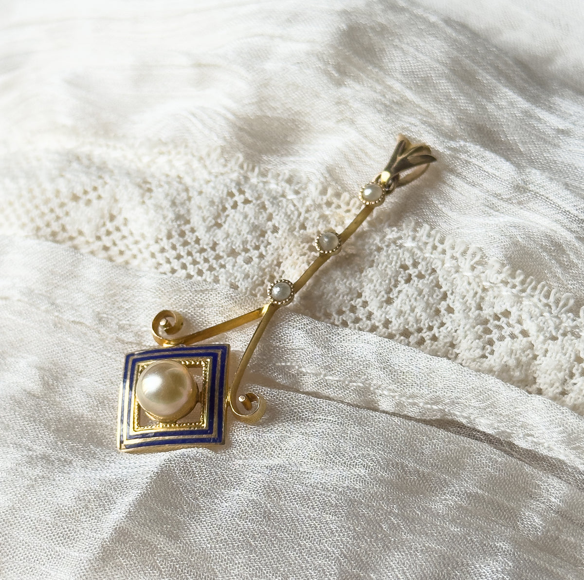 Art Deco pearl and seed pearl enamel lavalier pendant in 10K yellow gold, lying diagonally on white lace and gauzy fabric, its bail at top and cobalt blue enamel square at the lower end.