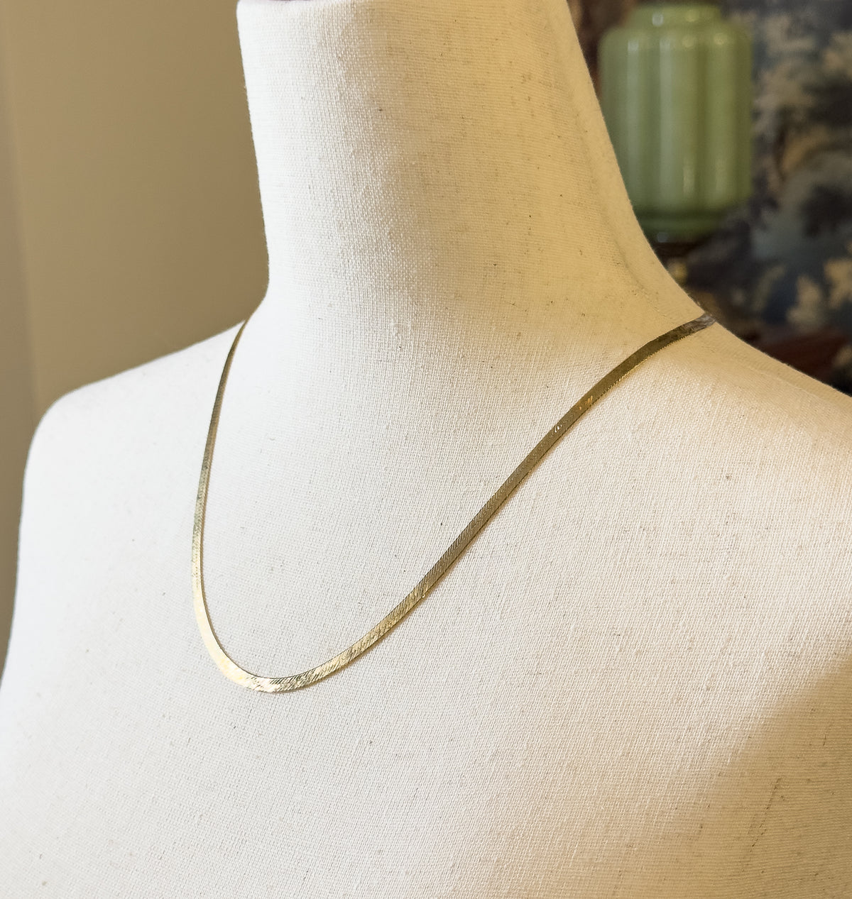 The flat herringbone chain shown at the neck on a cream fabric bust form, curving smoothly over the collarbone and lying close against the surface.