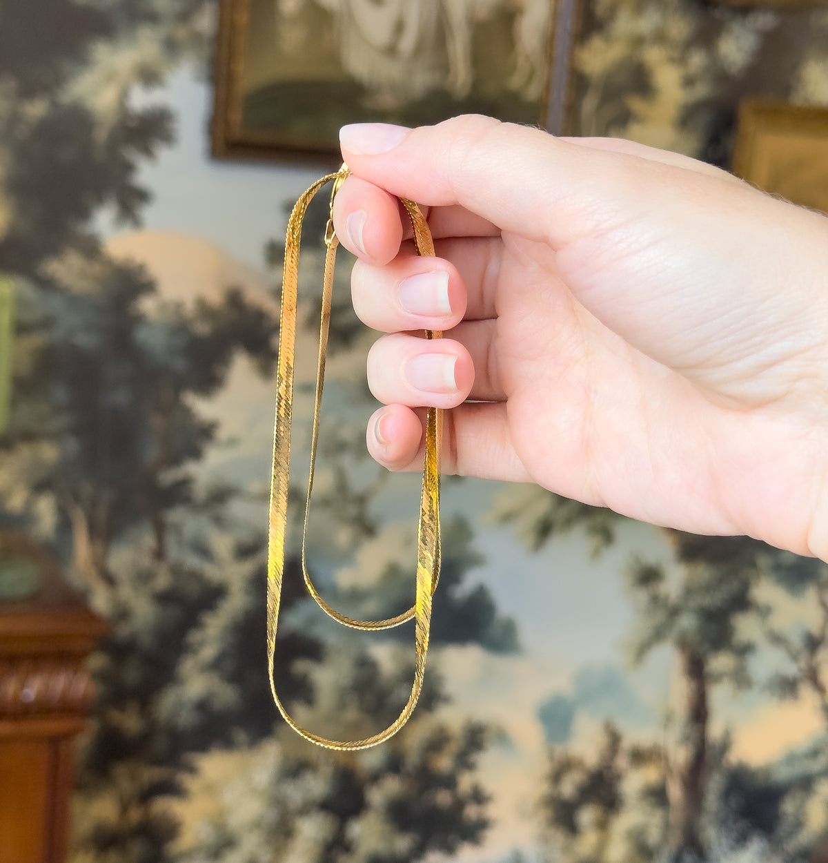 A herringbone chain necklace in 10K yellow gold, draped double and held between finger and thumb, the hand giving a sense of its slim, flat width.