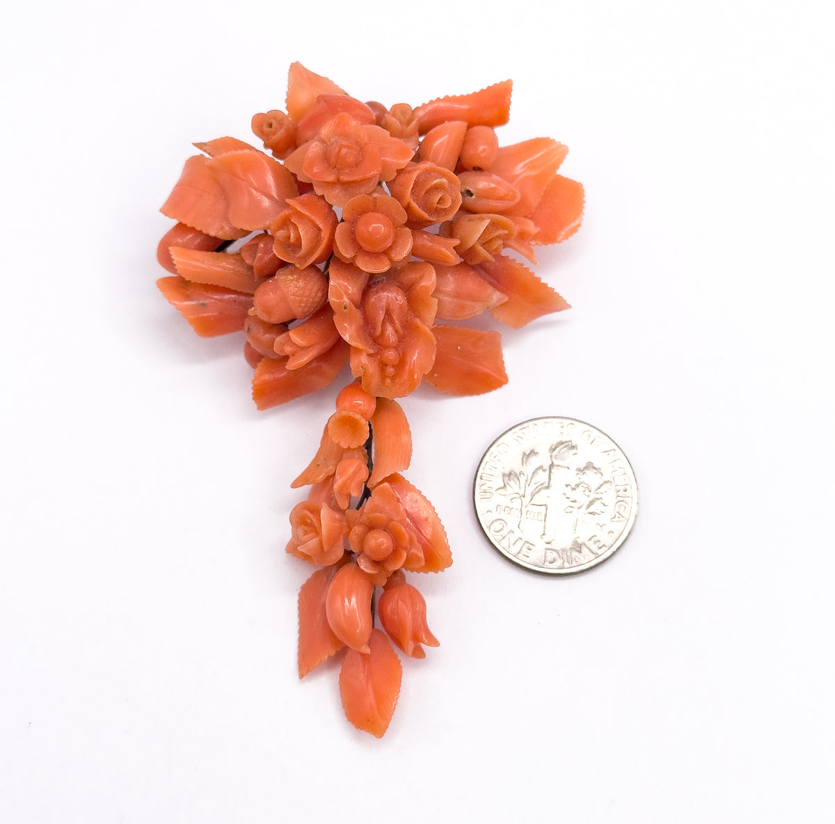 The carved coral pendant brooch face up beside a dime, the coin barely reaching across the trailing tail of leaves and buds, which shows the cluster stands several times the coin's width tall.