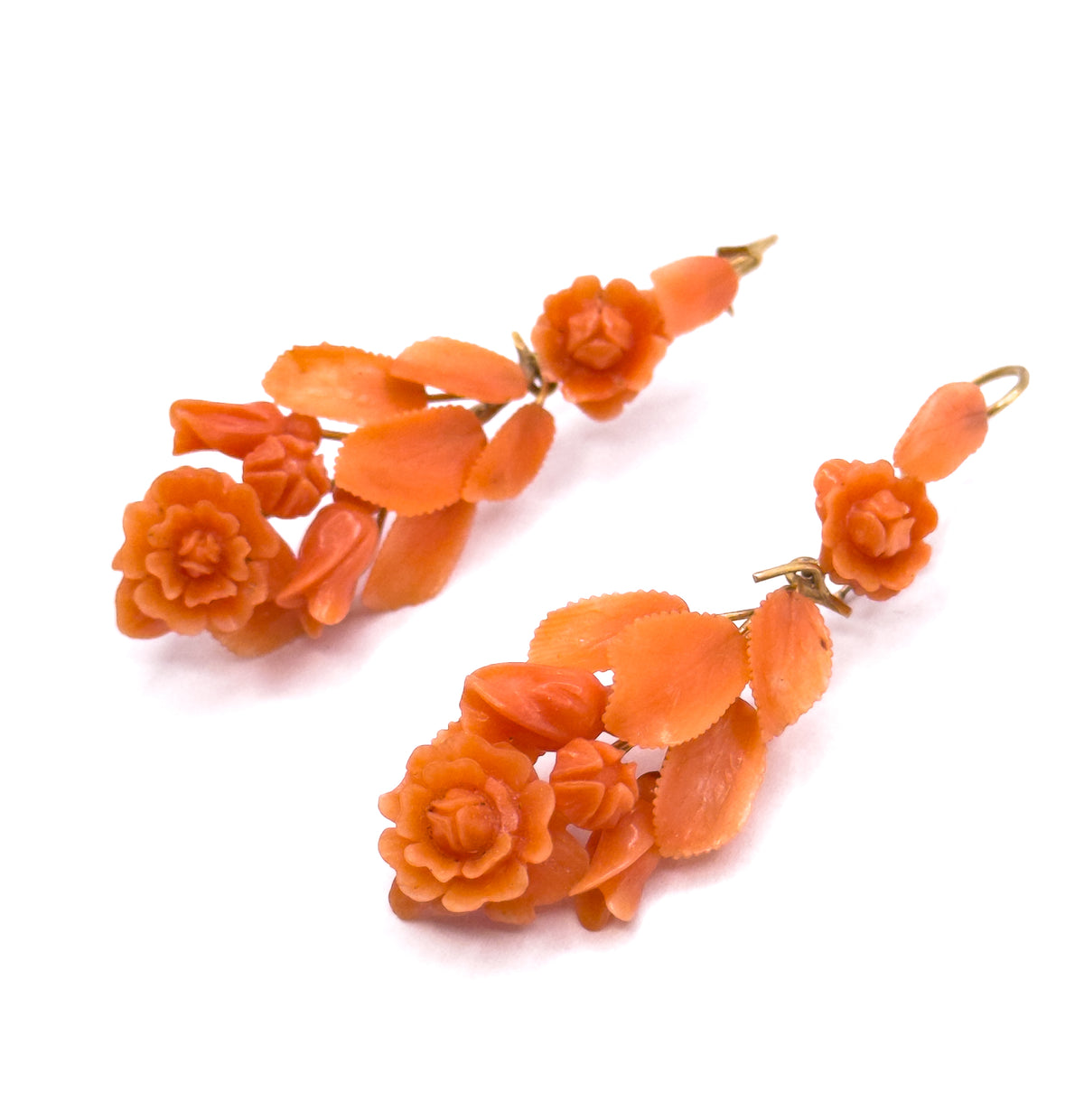 The pair lies flat and angled on white, showing the carved coral roses, buds and serrated leaves in dimensional relief along each yellow gold stem and its small loop top.