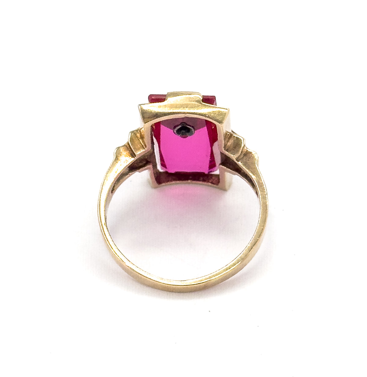Looking through the circle of the shank from behind, the open back of the setting shows the long red stone glowing pink from below with the small diamond visible at one end.