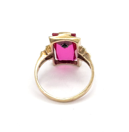 Looking through the circle of the shank from behind, the open back of the setting shows the long red stone glowing pink from below with the small diamond visible at one end.