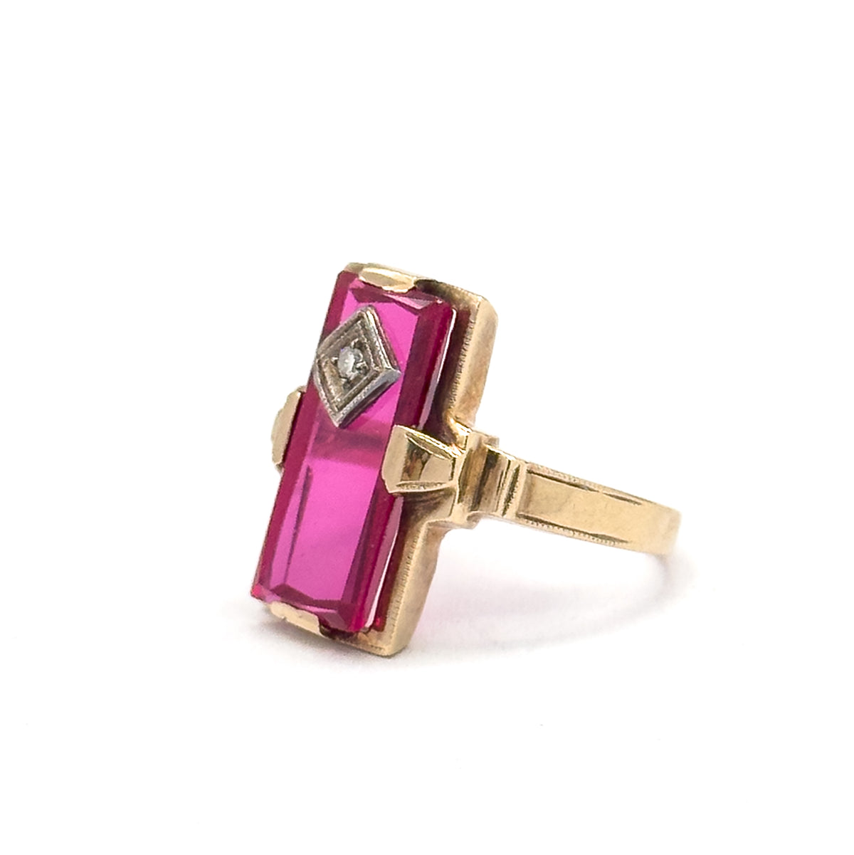 Angled three-quarter view from the front, the long red stone tilts toward the camera between its yellow gold corner prongs, the small diamond in its square bezel near the top, shank receding right.