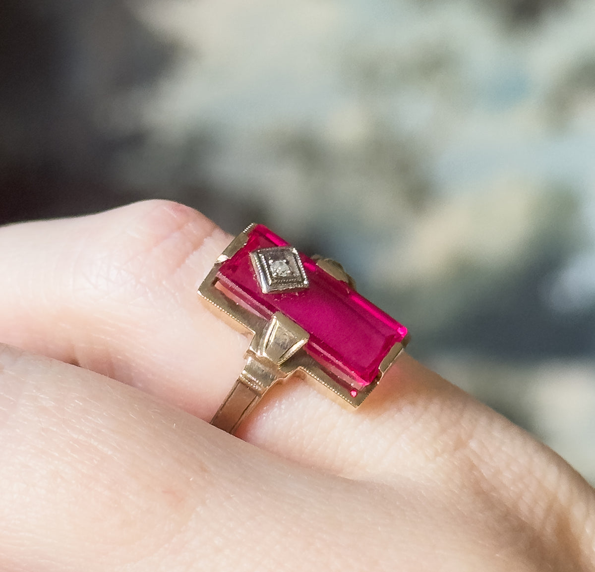 Retro lab-created ruby and diamond ring in yellow gold, shown on a finger, its elongated rectangular red stone set flat with a tiny diamond in a milgrain bezel at one end.