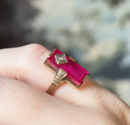 Retro lab-created ruby and diamond ring in yellow gold, shown on a finger, its elongated rectangular red stone set flat with a tiny diamond in a milgrain bezel at one end.