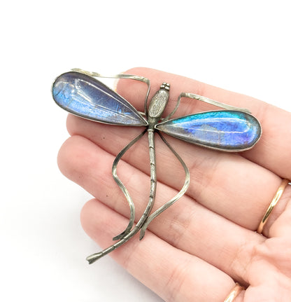 A sterling silver and base metal dragonfly brooch with blue and purple iridescent Morpho butterfly wings under clear domes, shown resting on an open palm for scale.