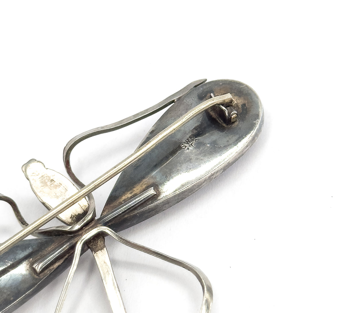 The reverse of the brooch, close in on the tail end, showing the long pin stem across the body, the hinged catch and small maker's marks struck into the darker patina.