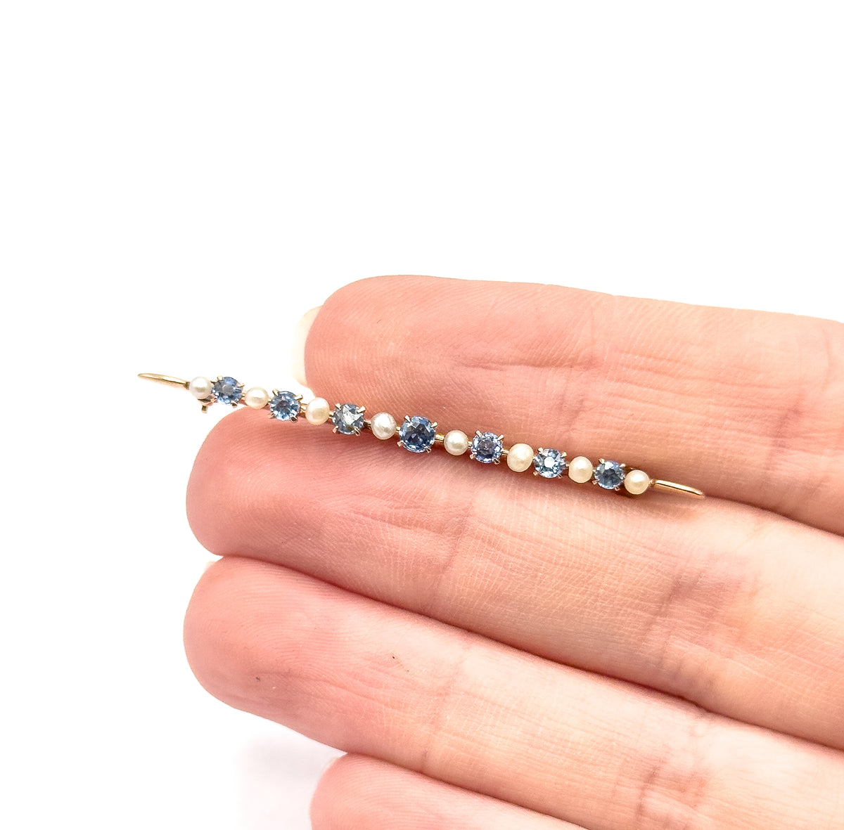 Edwardian sapphire and seed pearl bar brooch in 10K yellow gold, shown resting across the fingers of a hand for scale, its prong-set blue stones alternating with small pale pearls along the pin.