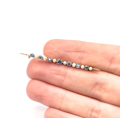 Edwardian sapphire and seed pearl bar brooch in 10K yellow gold, shown resting across the fingers of a hand for scale, its prong-set blue stones alternating with small pale pearls along the pin.