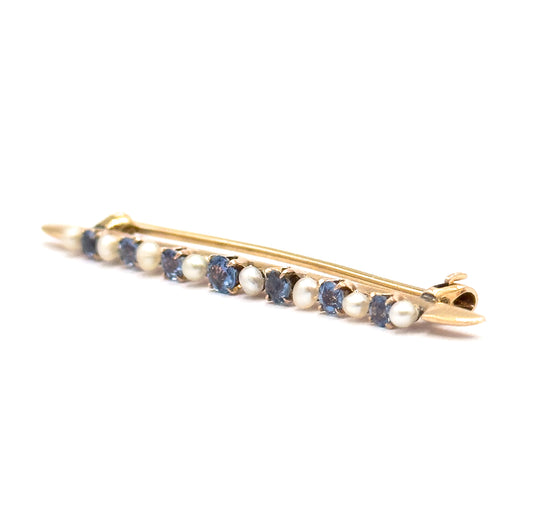 Low, near eye-level view along the bar with the catch end nearest the viewer, so the prong-set sapphires and seed pearls sit almost edge-on and the pin stem runs behind them.