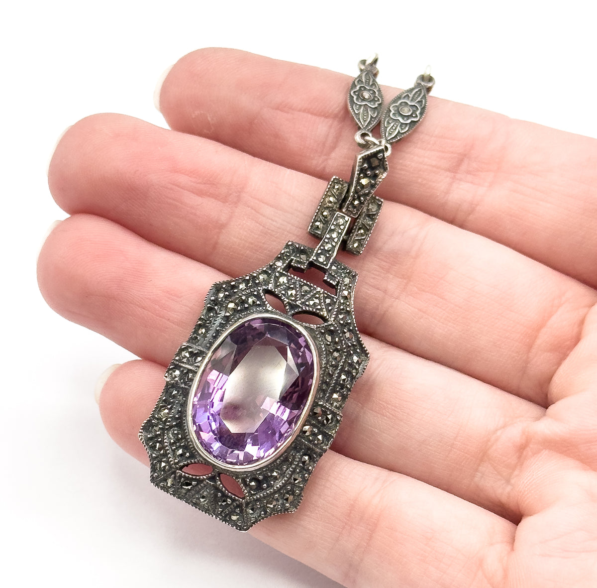 The necklace pendant shown resting across the fingers of a hand for scale, its oval amethyst face up in a milgrain bezel ringed with marcasite, with articulated links rising above.