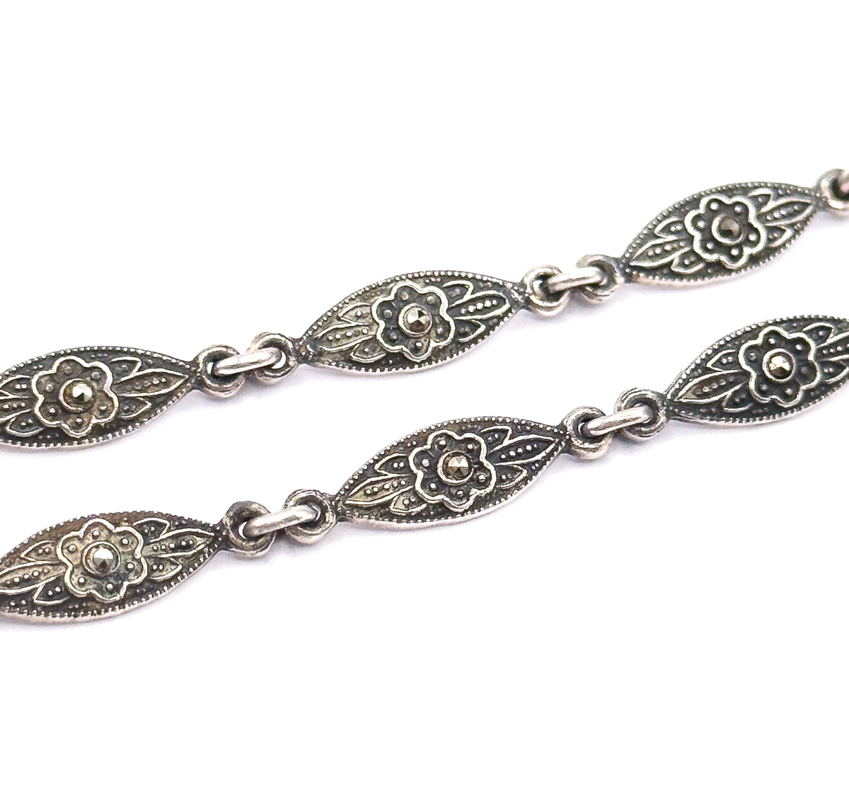 Close view of two strands of the necklace chain, each pointed-oval sterling silver link patterned with a flower centred on a marcasite within milgrain edging, joined by small round links.