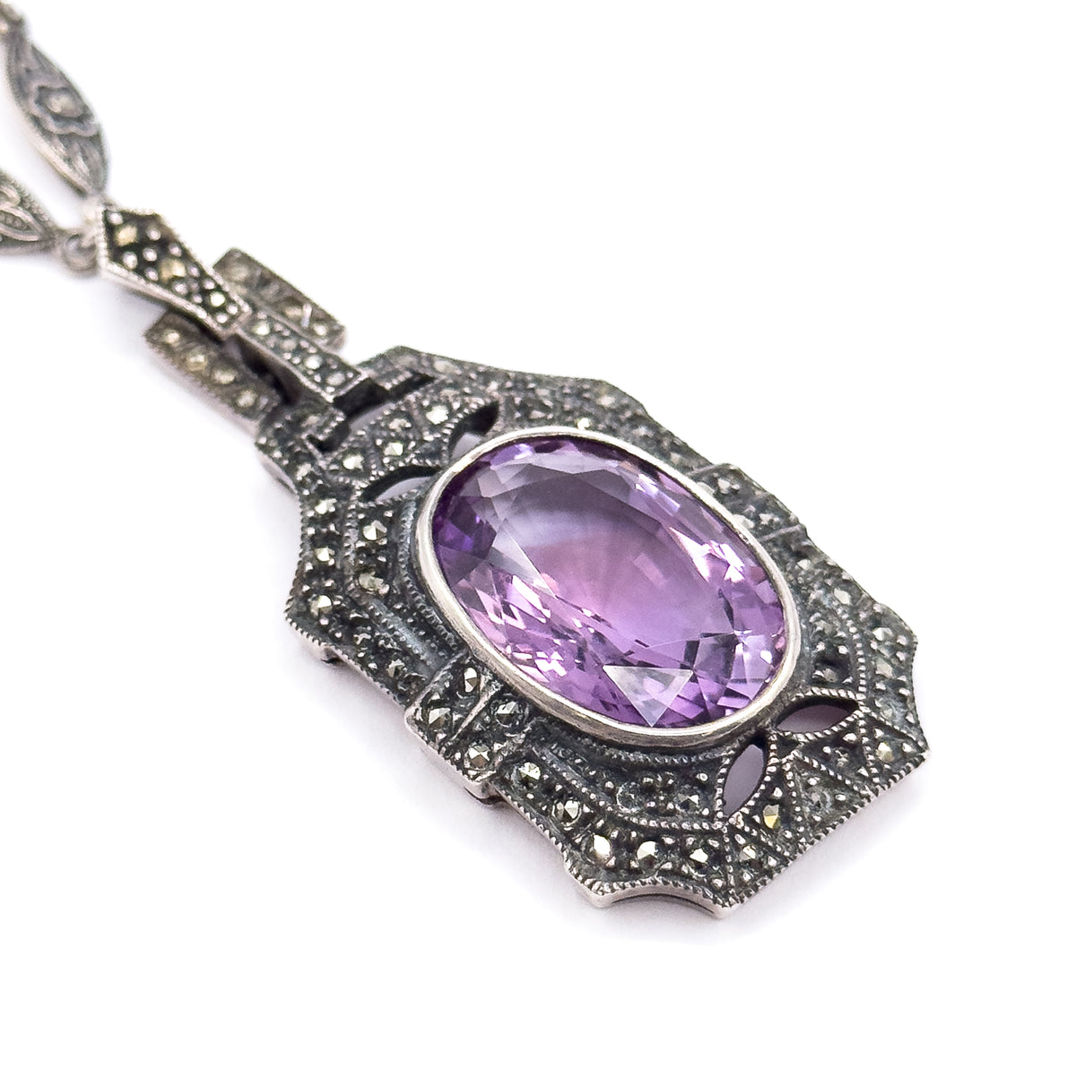 Angled three-quarter view of the pendant front, the oval amethyst in its milgrain bezel set in a stepped geometric frame paved with marcasite, with the articulated links and chain above.