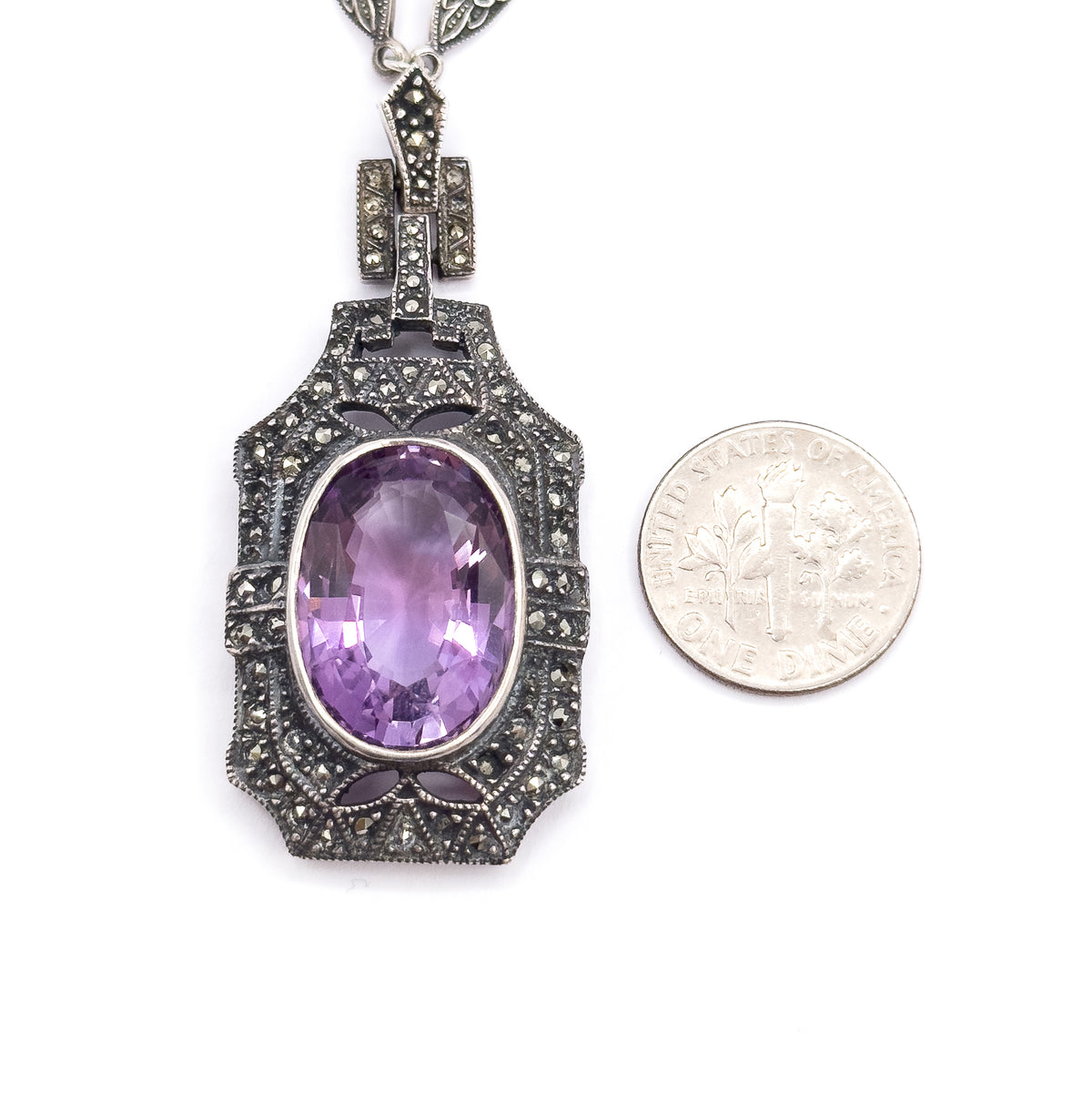 The pendant lies flat beside a dime for scale, the oval amethyst and its marcasite-paved frame standing noticeably taller and wider than the coin.