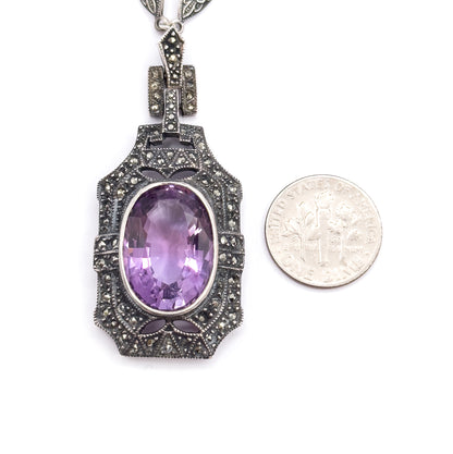 The pendant lies flat beside a dime for scale, the oval amethyst and its marcasite-paved frame standing noticeably taller and wider than the coin.