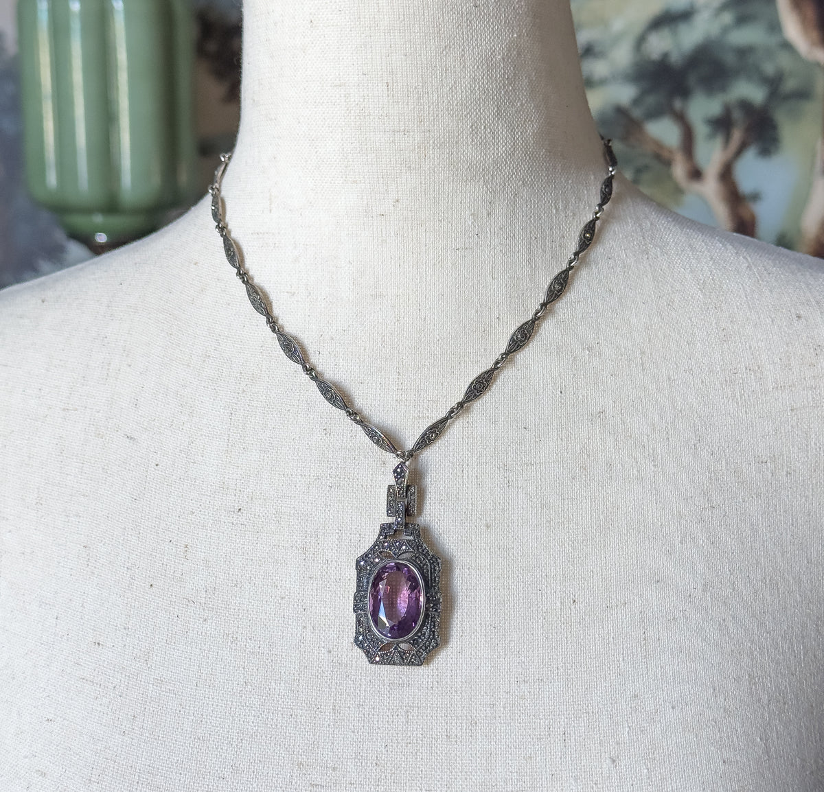 Sterling silver amethyst and marcasite necklace shown at the neck on a cloth mannequin, its oval purple stone set in a marcasite-edged rectangular pendant hung from a slim linked chain.