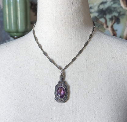 Sterling silver amethyst and marcasite necklace shown at the neck on a cloth mannequin, its oval purple stone set in a marcasite-edged rectangular pendant hung from a slim linked chain.