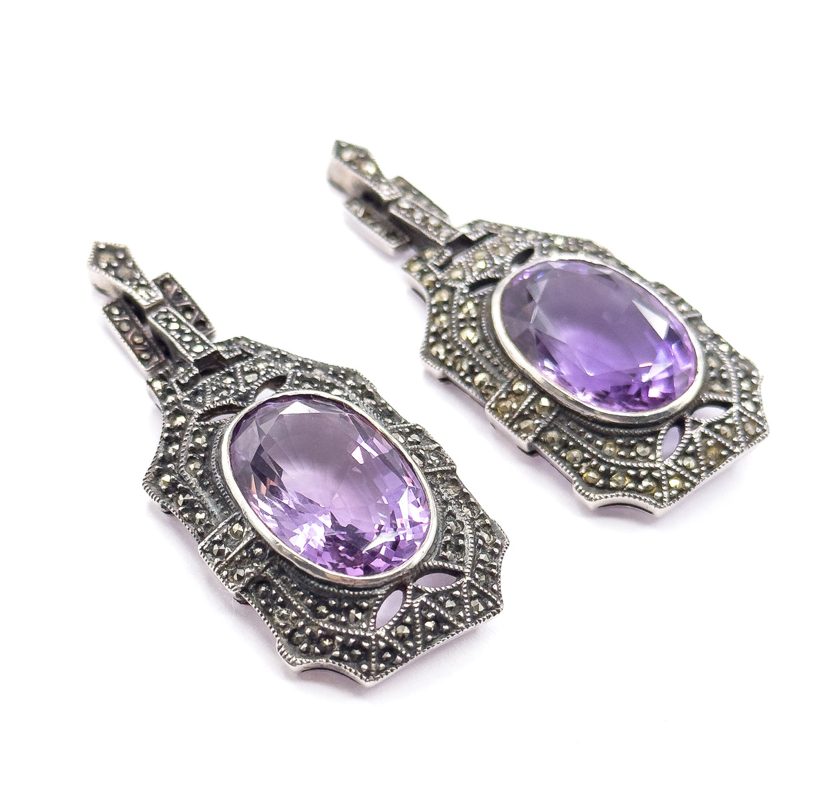 The pair of drop earrings lie side by side on white, each oval purple amethyst in a bezel framed by marcasite-set stepped panels with milgrain edges and small rectangular links above.