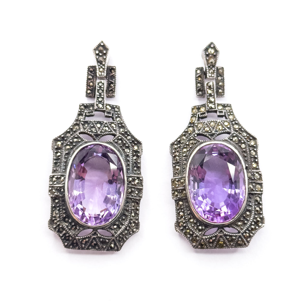 Close view of both earrings upright, showing the tapered marcasite tops, the open cut-out galleries and the stepped milgrain frames around each bezel-set oval amethyst.
