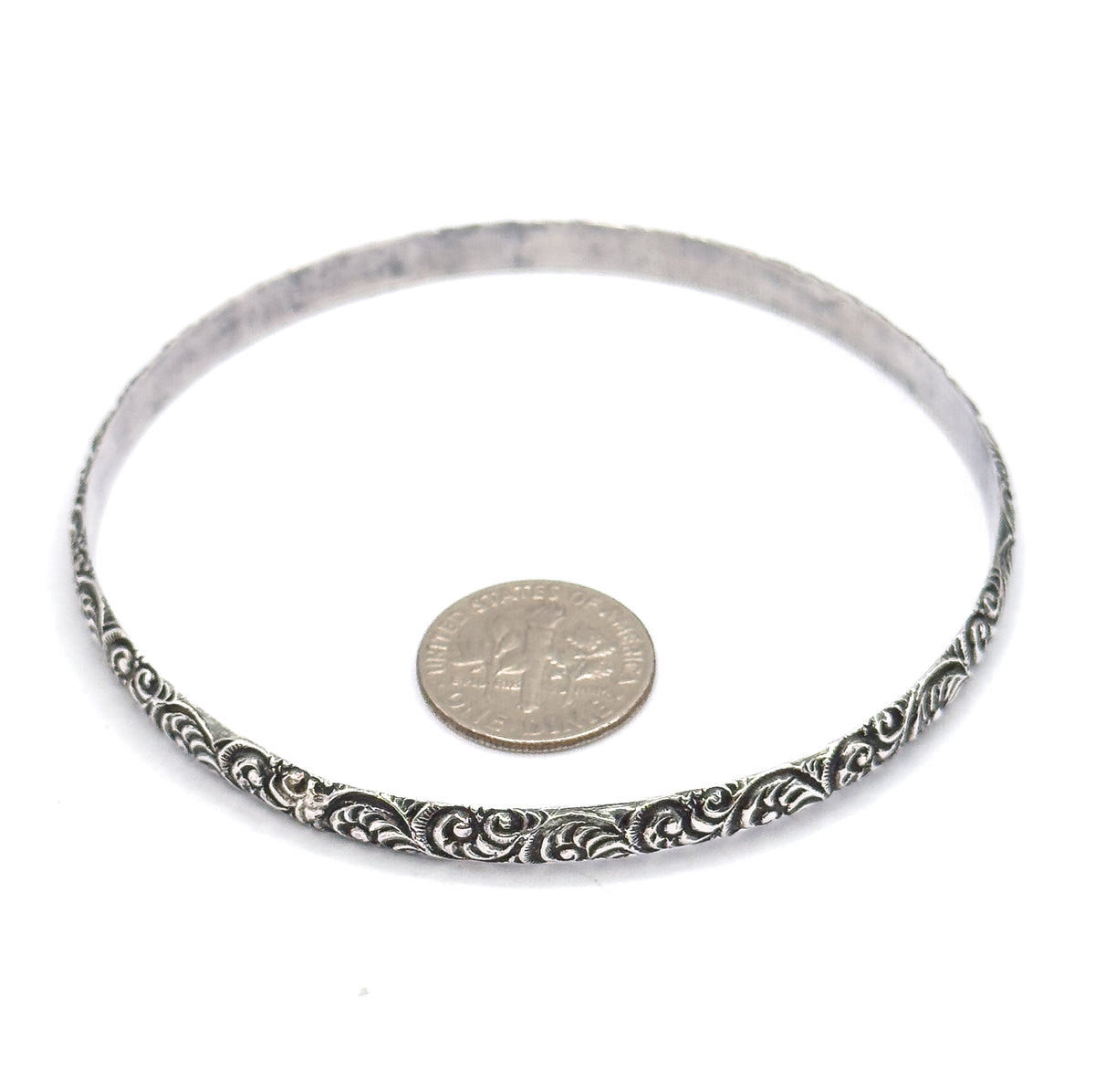 The bangle lies flat on a white background with a small coin set inside its oval opening for scale, the swirl pattern and darkened patina clearest along the front edge.