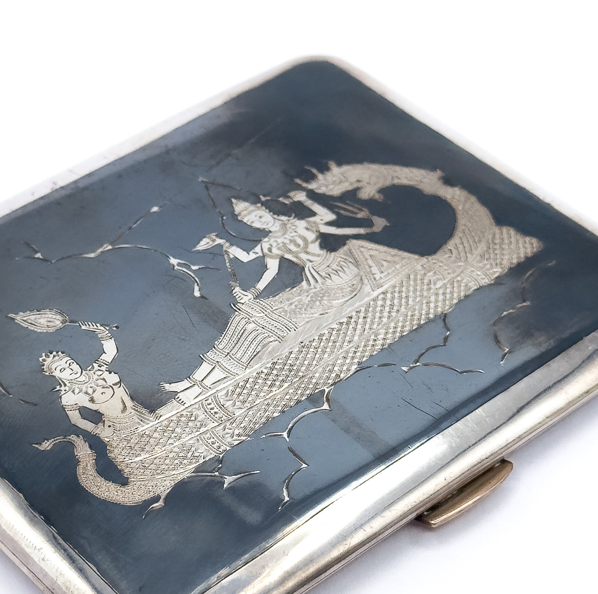 Tighter diagonal crop on the lid's engraving, where a crowned figure kneels raising a leaf-shaped form while a standing figure rides the scaled serpent boat, the clasp just visible at lower right.