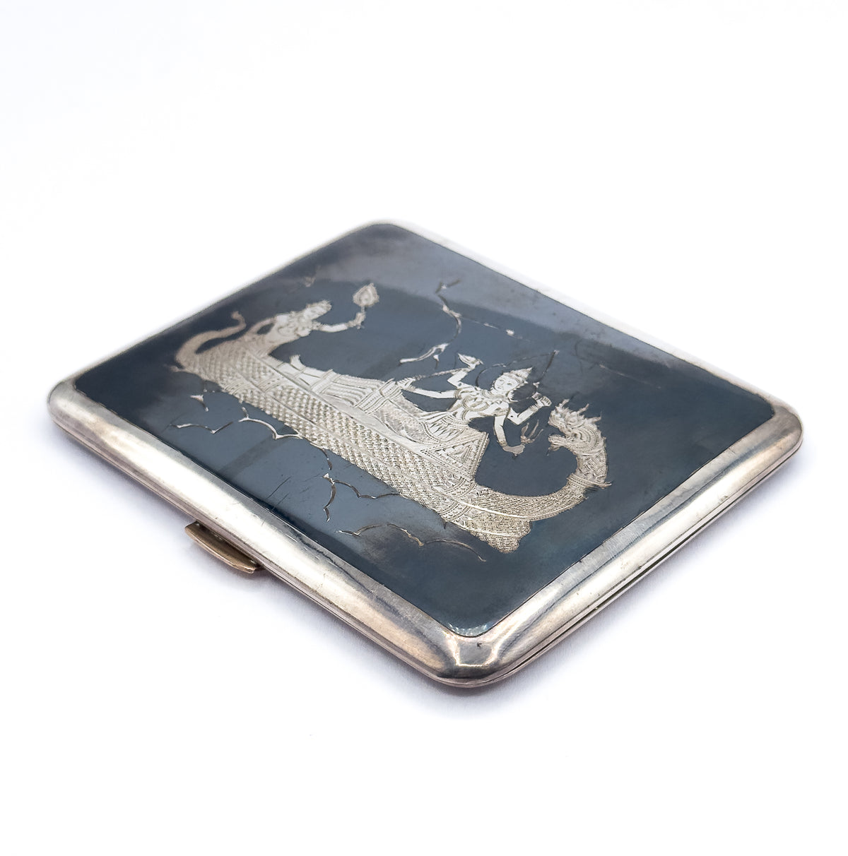 Closed and tilted on white, the lid's slate-black face is now toward the camera, engraved in Siam style with robed figures in a long boat, framed by the bright silver rim and clasp thumbpiece.