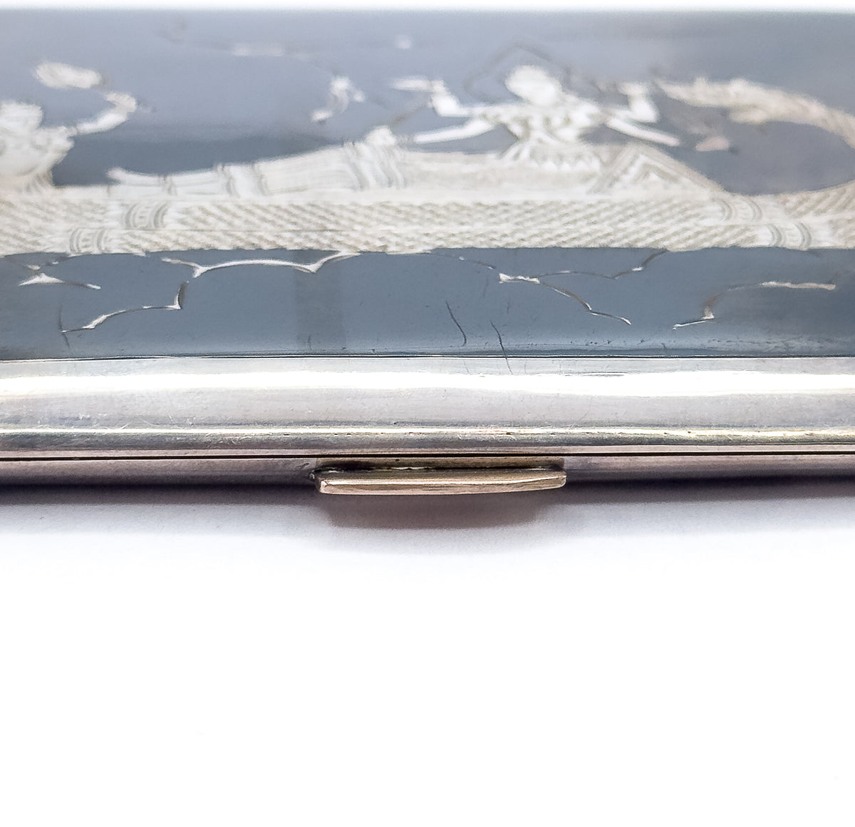 Close crop on the case's lower long edge, where the small clasp thumbpiece projects from the bright silver rim beneath the slate-black engraved panel and its band of figures.