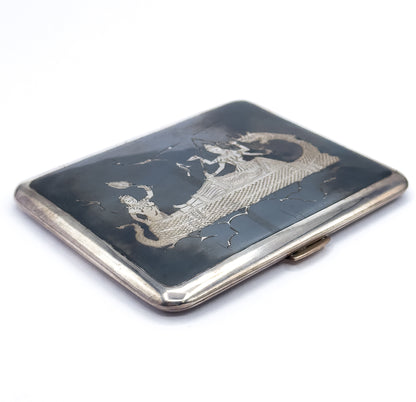 Angled from a lower corner, the closed case shows its full slate-black lid with engraved figures riding a long serpent-like creature, the bright silver rim running around to the clasp thumbpiece at right.