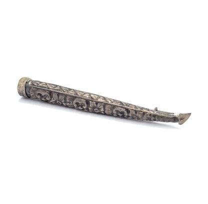 Laid on the diagonal with the collared rim at upper left, a band of small triangles edging the upper side above the scrolled repoussé, the pin stem emerging past the tapered tip at lower right.