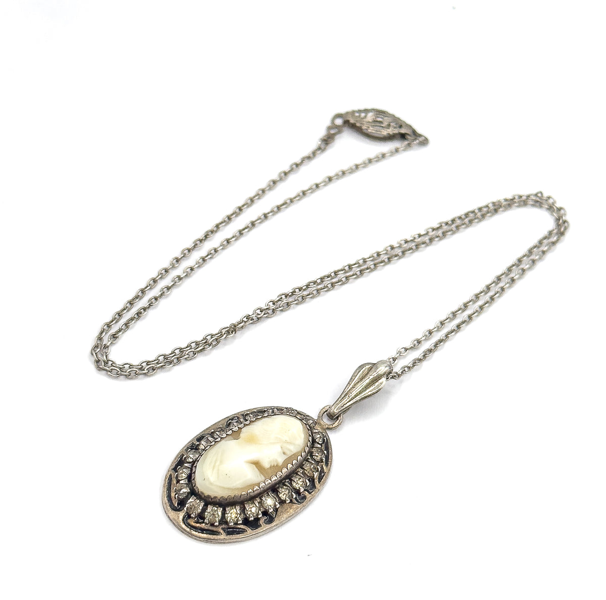 The front view shows the oval shell cameo's carved profile in its bezel, ringed by glass stones and enamel, with the fluted bail and chain looping up to the clasp.