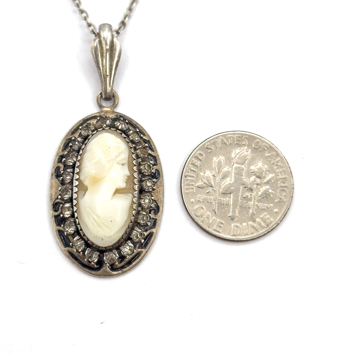 The pendant lies beside a dime for scale, the carved cameo and glass-stone halo standing about half again as tall as the coin, its chain rising off the top of the frame.