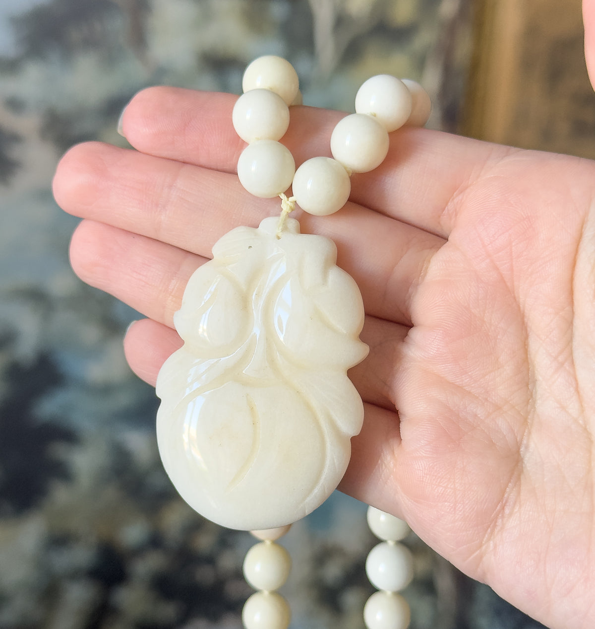 The carved gourd pendant rests across the fingers of an open hand, cord knot visible at its top, with the jade beads looping above and below the palm.