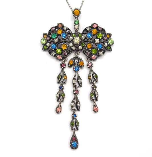 Edwardian paste bow necklace in dark-patinated sterling silver, shown front-on against white, its green, blue, pink and amber stones set through the bow and three tiers of leaf-shaped drops below.