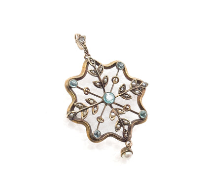 Front view tilted with the scalloped eight-lobed frame filling the frame, the bail at upper left, blue paste bezels on each arm, and the single pearl drop hanging from its cup at lower right.