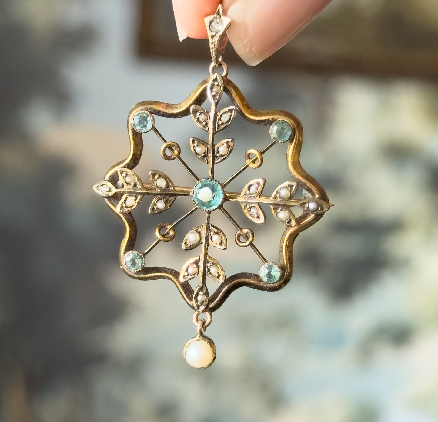 Edwardian starburst pendant in yellow gold with bezel-set icy blue paste stones and seed pearl leaves, held between fingertips by its bail, a small pearl drop swinging below.