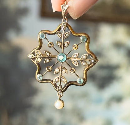 Edwardian starburst pendant in yellow gold with bezel-set icy blue paste stones and seed pearl leaves, held between fingertips by its bail, a small pearl drop swinging below.