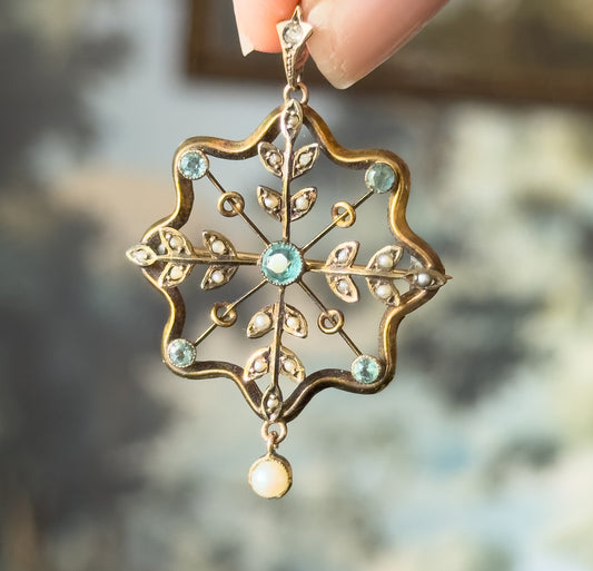 Edwardian starburst pendant in yellow gold with bezel-set icy blue paste stones and seed pearl leaves, held between fingertips by its bail, a small pearl drop swinging below.