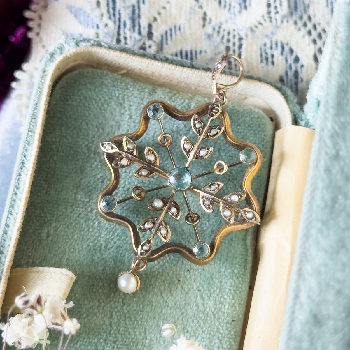 The pendant lies face-up in a pale green velvet ring box, its wavy gold frame, six radiating spokes, seed pearl leaves and hanging pearl drop seen from above.