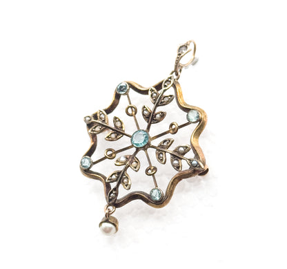 Angled front view on white with the central blue paste bezel sharpest, six radiating arms tipped by further bezels between seed pearl leaf sprays, bail at top right and pearl drop below.