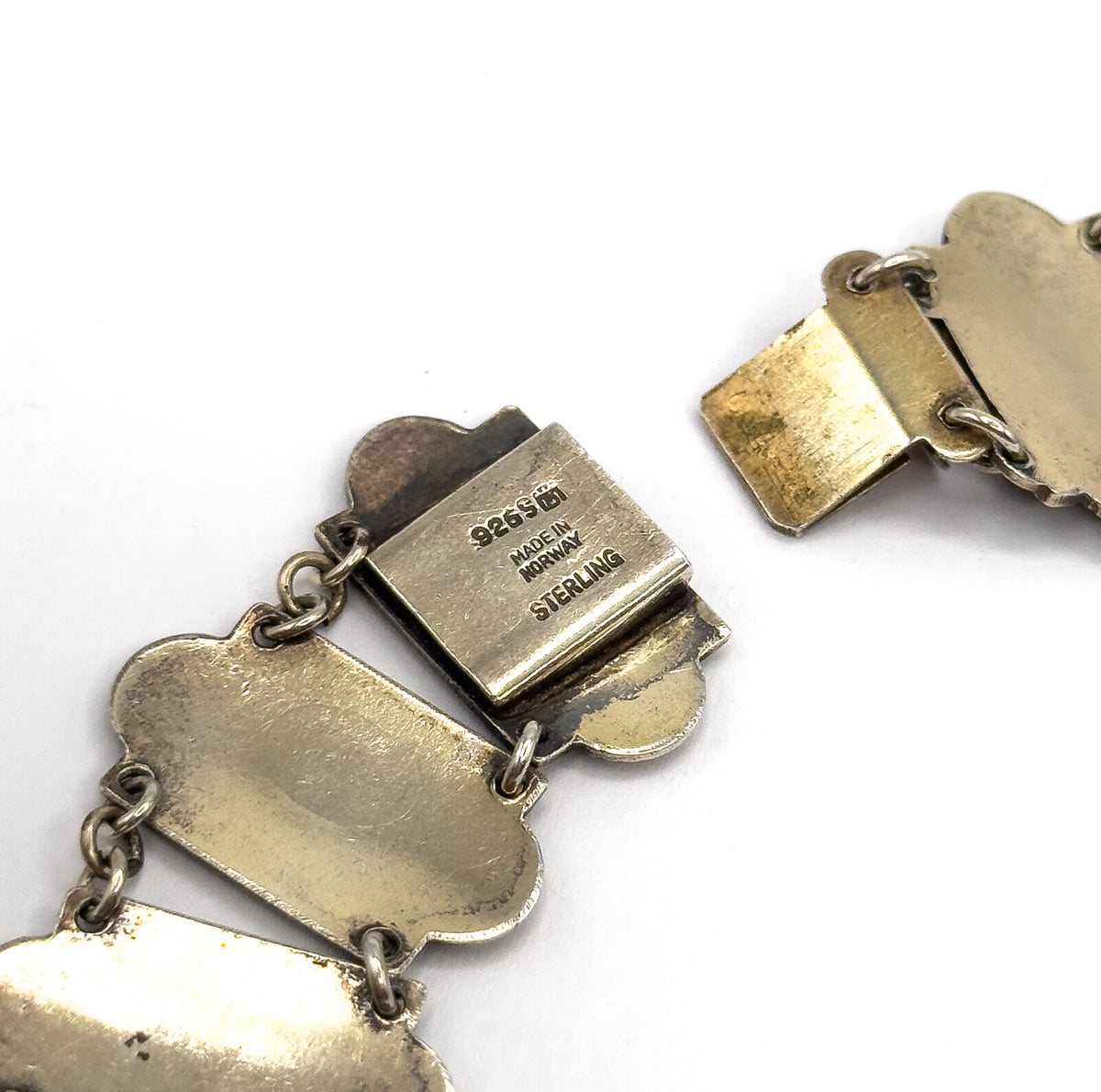 Close-up of the reverse of the open clasp, its flat box stamped with maker's markings and the flat tongue section lifted away at the right, with jump rings linking the plain link backs.