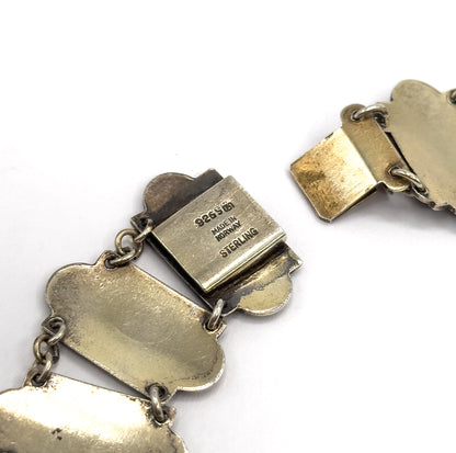 Close-up of the reverse of the open clasp, its flat box stamped with maker's markings and the flat tongue section lifted away at the right, with jump rings linking the plain link backs.