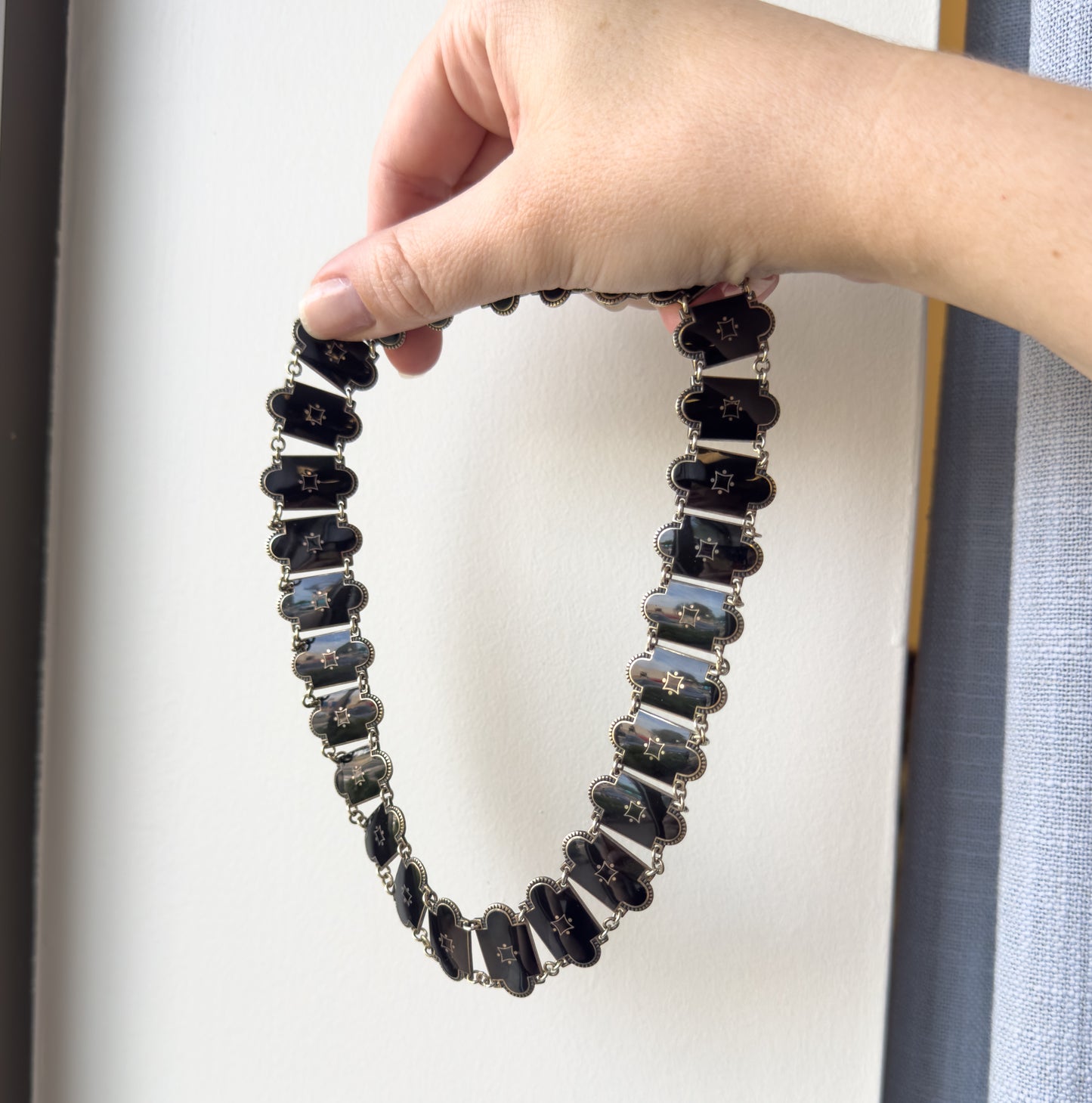 A black enamel station necklace in sterling silver, held up by a hand against a white wall so its ring of elongated links hangs in a full oval, giving a sense of its small size.