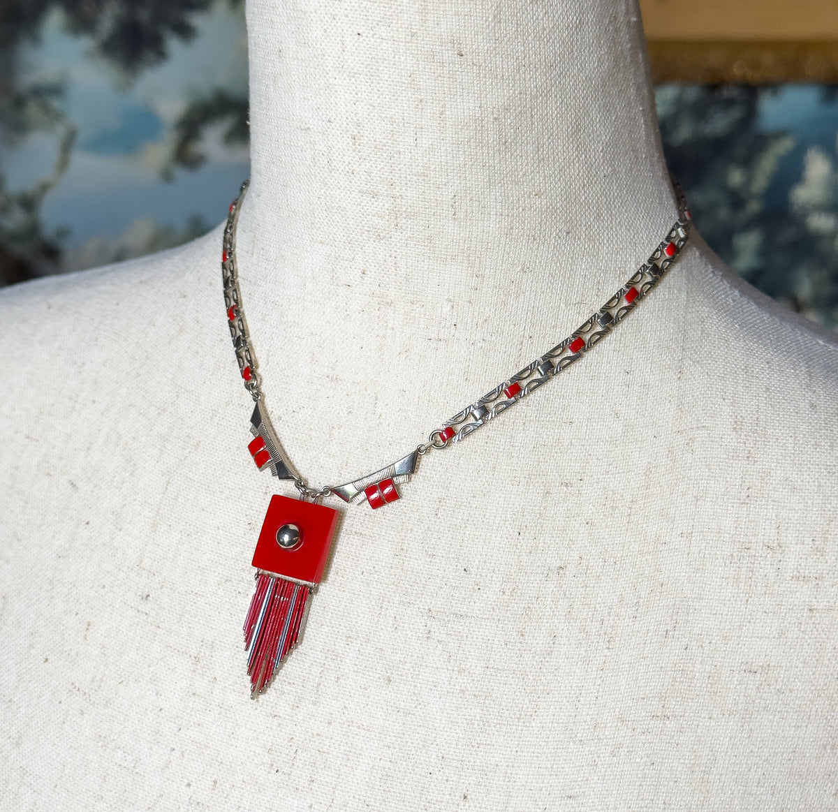 Closer angled view at the neck showing the light-coloured chain links dotted with small red enamel squares as they taper to the geometric plaque above the fringe.