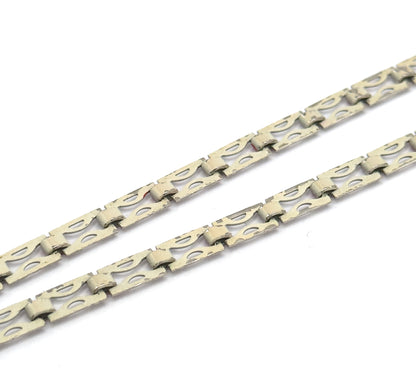 Macro close-up of two parallel runs of chain, each flat link pierced with a long teardrop opening and joined by small bar connectors, with a trace of red at one link.